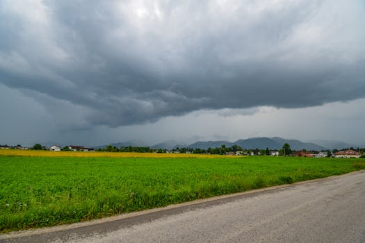 A storm brews over a lush green field with distant mountains and village houses.
