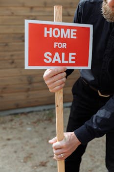 A person holding a red 'Home for Sale' sign outdoors in daytime, indicating real estate activity.